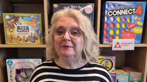 Kerry, a white haired woman in glasses, stands in front of shelf of toys in a shop, including Connect 4 and Toy Battle