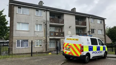 A three-storey block of flats at the end of a street in Bradford. There are balconies on the upper floors, and a police van is parked in front of the block.