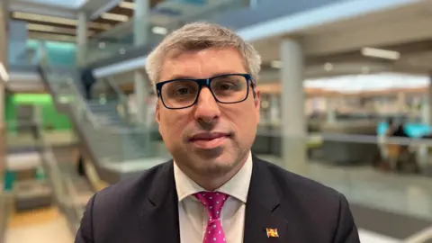 Mark Arnull is wearing a dark suit with a white shirt and purple patterned tie. He has a Northamptonshire flag pin. He is wearing dark glasses.