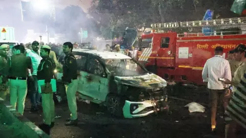 Indian police personnel inspect the scene of a car explosion. A white burnt out car can be seen, while a red fire engine is in the background