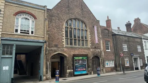 External view of St George's Guildhall in King's Lynn. The roof is a triangle shape and it is made of bricks of a wide range of shades of red, brown and black.
It has two vaulted doorways and signage at the front explaining what is happening at the building.