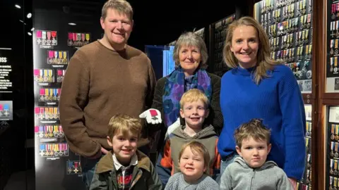 BBC/Claudia Robinson A family of four children and three adults stand among cases of medals in a museum exhibition.