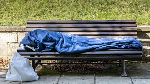 Getty A homeless person in a sleeping bag on a park bench. There is a bag beside the bench and green grass behind.