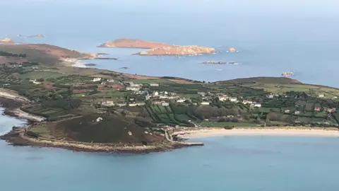An aerial photo of St Mary's on a sunny day. The island has fields and houses and a long white sand beach can be seen. There are further small islands beyond.