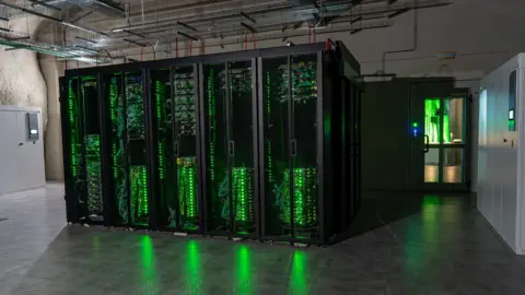 Trentino DataMine Green lights shine from a row of data servers in cabinets, reflecting on the high-gloss floor of the underground cavern. Metal trunking is attached to the ceiling above the cabinets, while to the right a door leads into a lighter room.