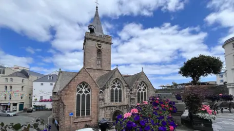 BBC A granite church with a slightly reddish hue, set among a shopping street. Purple and pink flowers in the foreground and a blue, cloudy sky.