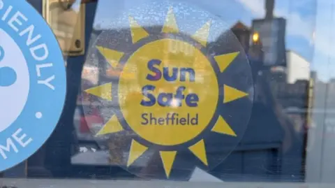LDRS A Sun Safe Sheffield sticker in the shape of a yellow sun displayed on the inside of a shop window.