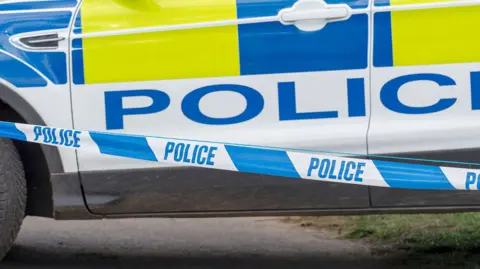A stock image of police tape and the side of a police car seen in the background. The tape is blue and white with the word police in capital letters on it several times.