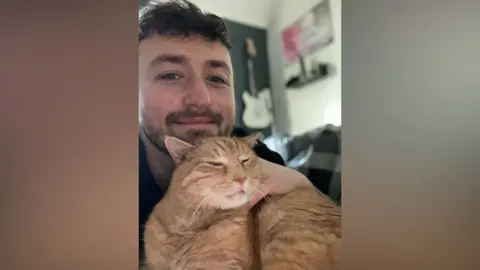 A selfie of a man with brown hair and a beard posing with a ginger cat.