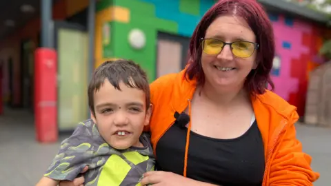 Shariqua Ahmed/BBC Logan, a boy with short hair, is wearing a grey and yellow T-shirt. Laura is a woman with red hair. She is wearing a black top and an orange hoodie. They are smiling for the camera.