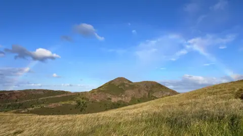 John Edward A grassy hillside rises toward a rugged green-and-brown hill beneath a bright blue sky. Faint rainbow arcs are visible on either side of the landscape, with scattered clouds adding depth to the scene.