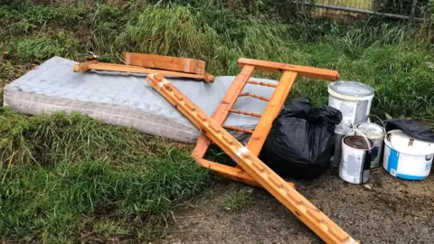 Gavin Cox A broken up pine bed and mattress with bin bags and old paint pots scattered on a patch of grass in the countryside.