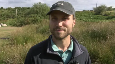Luke Engel is wearing a blue jacket and baseball cap and standing in front of moorland.