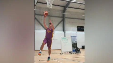 Tiverton Titans A basketball player throws an orange ball towards a basketball net. He is wearing a purple and yellow Tiverton Titans kit.