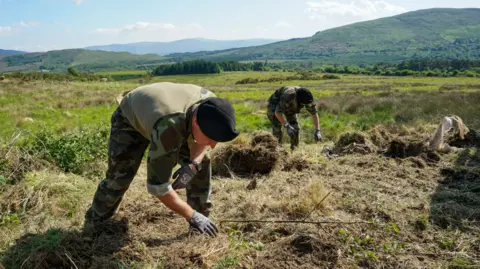 Two people wearing army clothing are digging in the ground with there hands. They are wearing gloves. In the background we can see a collection of hills and rugged countryside land. 
