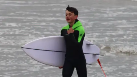 Neil Phillips Oscar has reached the shore after the rescue and he has his short surfboard tucked under his right arm and his left hand is tugging on the neck of his wetsuit.