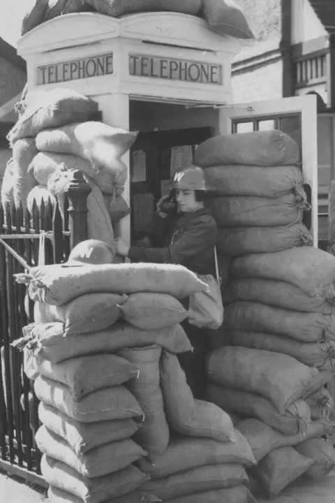 Keystone/Getty An archive, black-and-white, wartime photo showing a woman making a telephone call while standing in the entrance to a phone box, which is surrounded by stacks of sand bags on all sides and the roof. The woman is wearing a dark-coloured uniform and tin hat with a white "W" printed on it. 