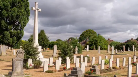 A photo of Gloucester Old Cemetery showing rows of gravestones and a tall stone cross.