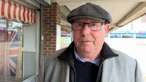 David Slim has a grey flat cap on, black glasses and a blond moustache. He is also wearing a grey coat over a blue jumper and blue shirt.