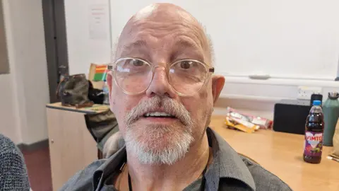 Phil Burdekin with glasses and receding grey hair and a grey beard wearing a grey shirt sits in a meeting room with a desk and whiteboard in the background.