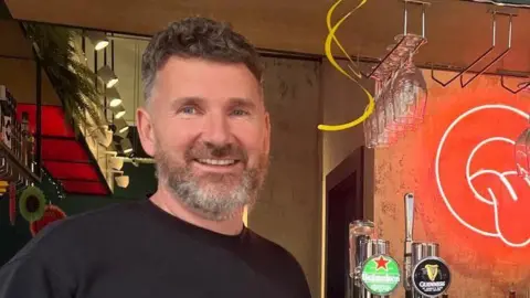 Arthur McAnerney. He has dark hair and grey beard, wearing a black t-shirt. He is standing by a bar with two beer drafts, a neon sign, and hanging glasses.