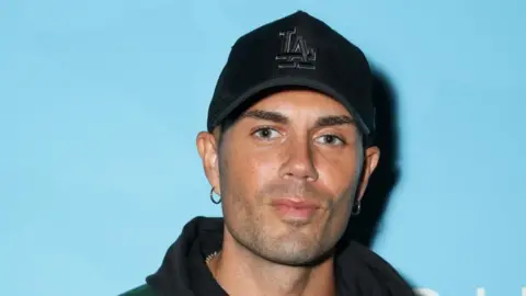 Getty Images A picture of Max George wearing a black baseball cap and a dark top while standing against a blue backdrop