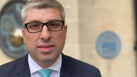 Mark Arnullwearing a black suit, lilac shirt and turquoise tie looking directly at the camera. He is also wearing black framed glasses.