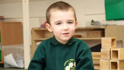 Lincoln Button - a five-year-old boy who is wearing a green jumper with a primary school logo on it. He is looking into the camera. 