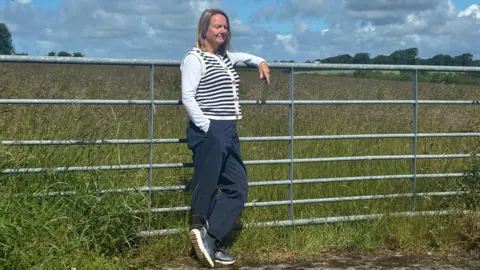 A woman stood leaning against a metal fence in a field. She has shoulder-length blonde hair and a white shirt with a black and white striped vest jumper on. She has dark blue trousers.