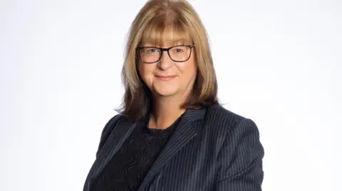 36 Crime A professional portrait of a woman with dark blonde shoulder length hair, she is wearing glasses and is smiling slightly at the camera. She is smartly dressed in a navy pinstripe jacket and a black top underneath.