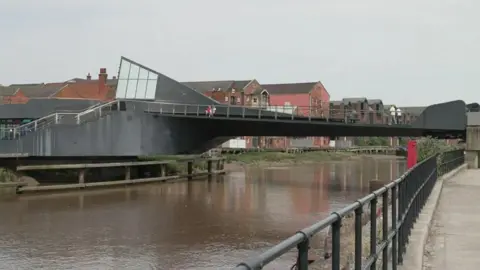The photo shows a black bridge that spans across a river. On the left hand side there are steps leading up to it. You can see people walking across it. Behind it are brick buildings that look like former industrial units. 