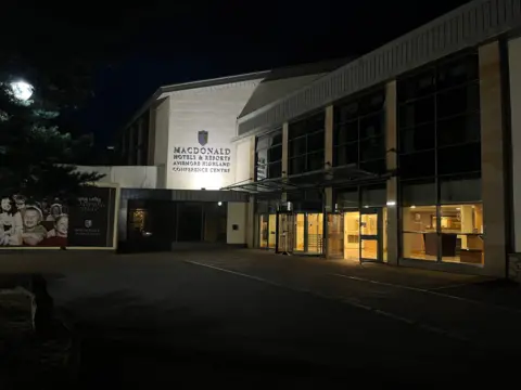 La entrada del Centro de Conferencias Macdonald Aviemore Highland se ilumina por la noche con luces que brillan a través de los grandes ventanales y puertas de cristal. El nombre del edificio se exhibe en la fachada principal, mientras que un letrero del Macdonald Aviemore Resort se encuentra junto a la entrada, en primer plano.