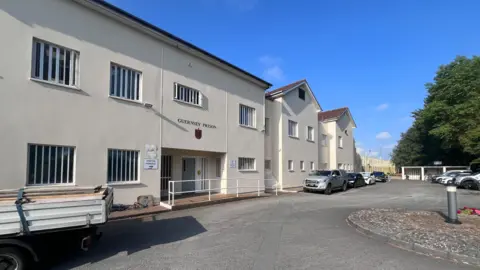 This is an exterior shot of Guernsey Prison. It is a cream building with a ground and first floor. There is a driveway with cars parked in front of it. There are tall trees behind them, and the sky is bright blue.