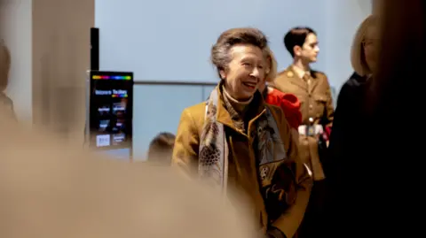 University of Gloucester Princess Anne smiles at a member of the public during her tour of the new campus building. She wears a brown coat and floral scarf.