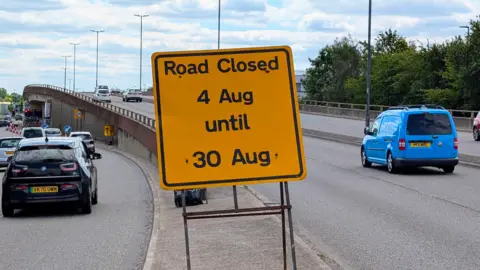 BBC A view of the flyover with cars driving over it. Several drive to the side of it. A yellow sign in the middle says "Road closed - 4 Aug until 30 Aug".