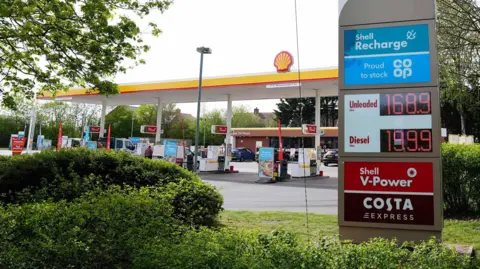 A Shell garage and forecourt. In the foreground is an electric sign which reads 168.9 for unleaded and 199.9 for unleaded fuel.