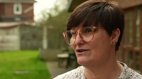 Sarah Crane, the minibus driver, with brown hair and wearing glasses. 