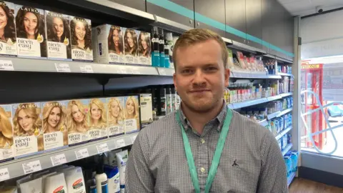 BBC Harry Pearson is standing in front of a shelf full of hair dye in the pharmacy. He has short blond/brown hair and is wearing a grey shirt and a lanyard.