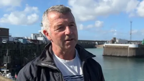 Stephen Viney pictured at the harbour. The sea and the pier are visible behind him. He has a black jacket on and short grey hair. 