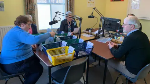 Four people, two men and two women, sit around desks with newspapers and baskets of newspaper cuttings in front of them. Three of them have microphones in front of them also. A woman sits in the background listening in.