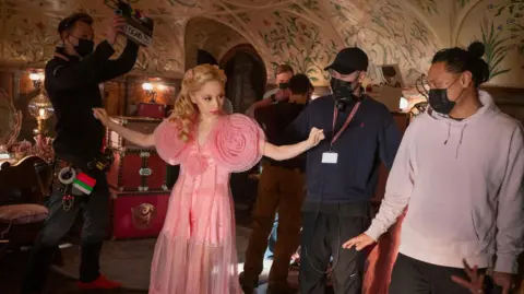 Universal Pictures/Sky Studios Elstree An actor wearing a frilly pink dress stands with her arms out, surrounded by film crew, one has a clapper board. The crew are wearing masks