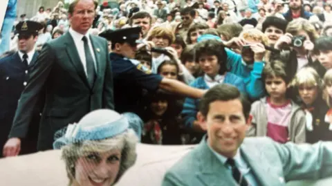 University Hospital Southampton Princess Diana and then-Prince Charles sit in a white car in the foreground of the photo. Prince Charles is wearing a grey suit and Princess DIana has on a blue fascinator. In the background, a man in a dark suit can be seen - this is Allan Peters.