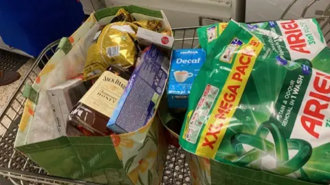 CAMBRIDGESHIRE POLICE Two large vinyl shopping bags seen from a bird's eye view in a shopping trolley. In the bag on the left, there is a bottle of Jack Daniels whiskey, as well as confectionery items from brands including Milka and Mikado. In the bag on the right, there are Ariel branded laundry products and a box of Lavazza tea or coffee. 
