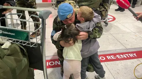A soldier in a camouflage uniform and blue hugs a young girl in a white top and pink trousers and a small boy in a grey jumper in the airport.