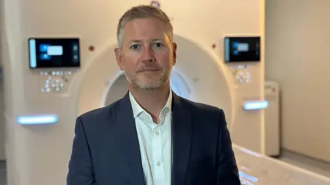 A man, with grey hair, a beard and a moustache, wearing a suit jacket and white shirt, stands in front of an MRI scanner with a neutral expression on his face. A cylindrical scanner, with screens on either side, can be seen, slightly out of focus, behind him.