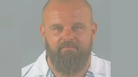 Hampshire and Isle of Wight Constabulary Lee James' custody picture. He has a shaved head and a medium-length beard with grey hairs in it, and looks at the camera wearing a blue and white shirt.