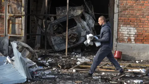 Reuters A person walks down a street with rubble and mess on the floor with a burnt out car inside a garage on the wall, depicting a destroyed car mechanics garage, in Dnipro on Tuesday.
