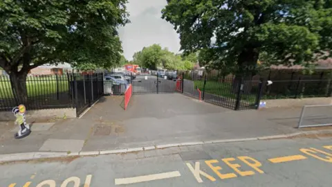 Google The words school keep clear are in yellow on the road at the bottom of the photo. Black railings, vehicles and buildings are behind the pavement in the foreground in this generic image.
