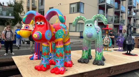 Three colourful Wallace sculptures. They depict dogs. One is red with multicoloured patterns, one is green with floral patterns and one is yellow with blue and red striped legs.