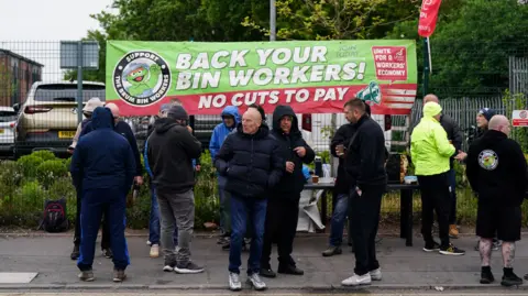 PA Media A gathering of men in dark clothing in front of a hedge with a large red banner which reads back your bin workers